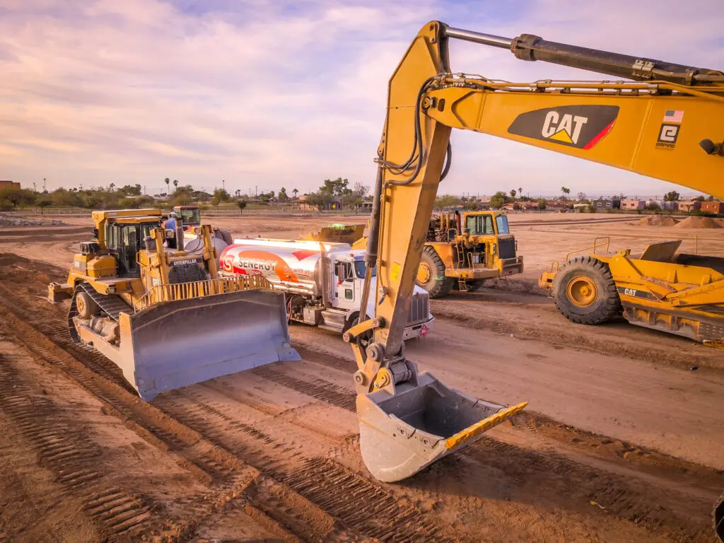 A Senergy Petroleum fuel truck refueling heavy construction equipment at a worksite. The scene includes a large excavator arm in the foreground, along with a bulldozer and other machinery on a dirt surface under a cloudy sky.