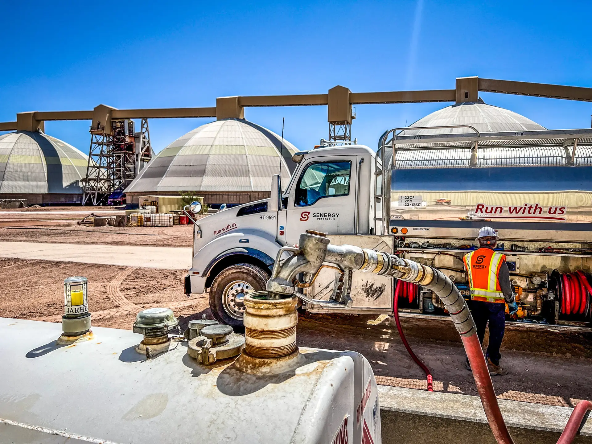 A Senergy Petroleum fuel truck parked at an industrial site with large storage domes in the background. A worker wearing a high-visibility vest and hard hat is operating equipment beside the truck, preparing to dispense fuel."